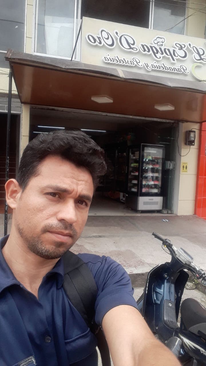Hombre con barba tomándose una selfie frente a una panadería con una moto estacionada.